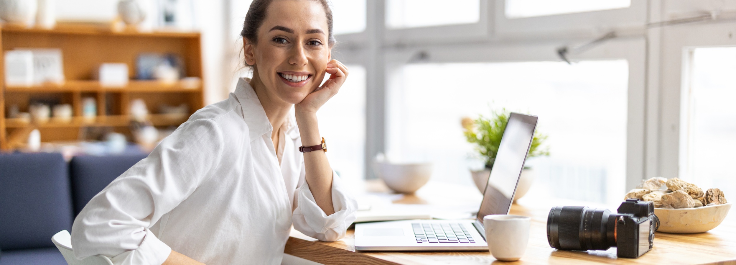 Eine lächelnde Frau sitzt an einem Schreibtisch mit einem Laptop, einer Tasse, einer Kamera und einer Schale mit Dekorationen in einem hellen, modernen Raum.

Das Bild zeigt eine junge Frau mit braunen Haaren, die zu einem Dutt hochgesteckt sind, die in einer weißen Bluse an einem hellen Holztisch sitzt und in die Kamera lächelt, wobei ihre linke Hand ihr Kinn stützt. Auf dem Tisch vor ihr befindet sich ein geöffneter Laptop, eine weiße Kaffeetasse, eine schwarze Kamera mit Objektiv, ein Notizbuch und eine Schale mit dekorativen Steinen oder Holzelementen. Hinter dem Tisch ist ein großes Fenster zu sehen, das viel Tageslicht hereinlässt, und eine Pflanze in einem Topf. Im Hintergrund ist ein Teil eines Bücherregals mit verschiedenen dekorativen Gegenständen und ein blaues Sofa zu erkennen. Die Atmosphäre ist hell, freundlich und einladend, was auf ein entspanntes und produktives Arbeitsumfeld hindeutet. Die Stimmung des Bildes ist positiv und optimistisch.
