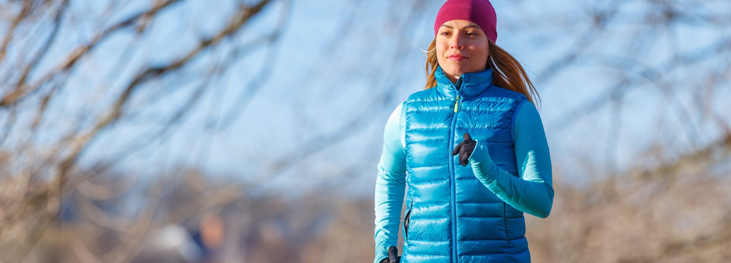 Das Bild zeigt eine Frau mittleren Alters, die im Freien joggt und dabei sportliche Kleidung trägt.

Im Detail zeigt das Bild eine Frau, die sich beim Joggen oder Laufen im Freien befindet. Sie trägt eine purpurrote Mütze, eine türkisfarbene, gesteppte Weste und ein langärmeliges Oberteil in einem ähnlichen Farbton. Ihre Hände sind mit schwarzen Handschuhen bedeckt. Die Frau scheint sich in einem Park oder einer ähnlichen Grünanlage zu befinden, da im Hintergrund Bäume und ein blauer Himmel erkennbar sind. Die Äste der Bäume sind kahl, was auf eine kühlere Jahreszeit hindeutet. Das Licht ist hell und sonnig, was eine positive und dynamische Atmosphäre erzeugt. Die Frau wirkt fokussiert und entschlossen, was die Stimmung von Aktivität und Gesundheit unterstreicht.