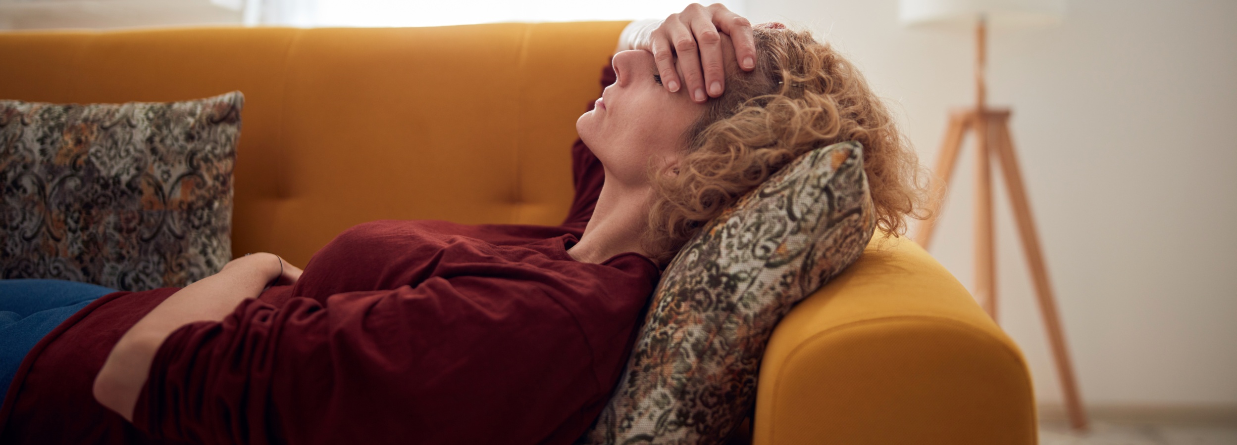 Eine Frau liegt verzweifelt auf einem Sofa und hält sich die Stirn.

Auf dem Bild ist eine Frau zu sehen, die auf einem leuchtend gelben Sofa liegt. Sie hat lockiges blondes Haar und trägt ein dunkelrotes Oberteil mit langen Ärmeln über einem blauen Kleidungsstück. Ihre rechte Hand liegt auf ihrer Stirn, und ihr Kopf ist leicht nach hinten geneigt, was auf Erschöpfung oder Verzweiflung hindeutet. Sie ist von zwei gemusterten Kissen umgeben, eines hinter ihrem Kopf und eines unter ihrem Arm. Eines der Kissen hat ein auffälliges Muster in Grün-, Braun- und Beigetönen. Der Hintergrund ist unscharf, zeigt aber ein weißes Wandobjekt, das wie eine Stehlampe aussieht, und eine helle Wand. Die Beleuchtung ist weich und diffus, was die melancholische und nachdenkliche Atmosphäre des Bildes verstärkt. Die Stimmung ist insgesamt ruhig und intim, aber durch die Körperhaltung der Frau wird eine tiefe emotionale Belastung oder ein Kampf mit inneren Schwierigkeiten vermittelt.