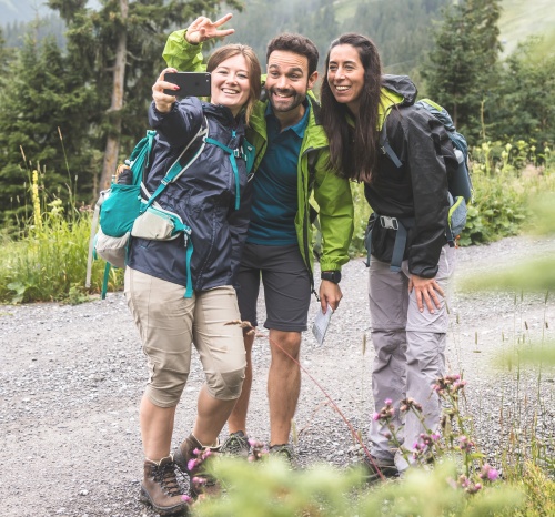 Das Bild zeigt drei fröhliche Wanderer, die auf einem bergigen Wanderweg ein Selfie machen und die gemeinsame Zeit in der Natur geniessen.

Im Bild sind drei Personen zu sehen, zwei Frauen und ein Mann, die sich auf einem Schotterweg in einer bergigen Landschaft befinden. Sie sind offensichtlich beim Wandern, denn sie tragen Rucksäcke und Wanderkleidung. Die Person ganz links, eine Frau mit blonden Haaren, hält ein Smartphone und macht ein Selfie mit ihren beiden Begleitern. Sie trägt eine blaue Regenjacke, beige 3/4-Hosen und braune Wanderschuhe. An ihrem Rucksack ist eine türkise Wasserflasche befestigt. Sie lächelt breit und macht eine V-Geste mit zwei Fingern.

Neben ihr steht ein Mann mit dunklem Haar und Bart, der eine grüne Regenjacke über einem blauen T-Shirt und dunklen Shorts trägt. Er lächelt ebenfalls und schaut in die Kamera. Er hält eine kleine Karte oder ein Notizbuch in der Hand.

Ganz rechts ist eine Frau mit dunklem Haar zu sehen, die eine schwarze Regenjacke über einer grauen Wanderhose trägt. Auch sie lächelt und scheint in das Selfie involviert zu sein. Sie trägt einen schwarzen Rucksack.

Der Hintergrund des Bildes zeigt eine üppig grüne Berglandschaft mit vielen Bäumen und sanften Hügeln. Der Himmel ist bewölkt, was auf ein kühleres Wetter hindeutet, passend zur Kleidung der Wanderer. Im Vordergrund sind vereinzelt blühende lila Disteln zu sehen, die dem Bild eine natürliche und leicht wilde Note verleihen.

Die Atmosphäre des Bildes ist sehr positiv, fröhlich und unbeschwert. Die lächelnden Gesichter und die Selfie-Pose vermitteln ein Gefühl von Freundschaft, gemeinsamer Freude und Abenteuerlust. Die Naturkulisse trägt zu einer friedlichen und erfrischenden Stimmung bei. Es ist offensichtlich, dass die drei Personen ihre gemeinsame Wanderung und die Natur geniessen.