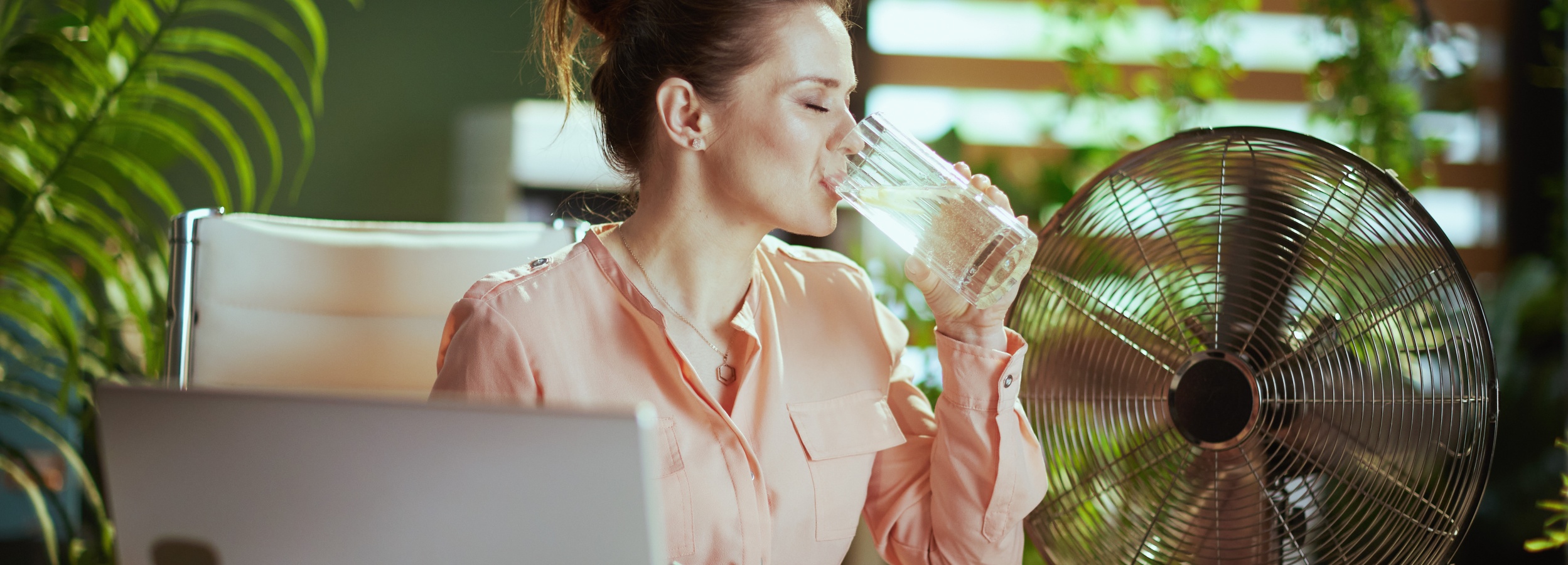 Junge Frau mit rosafarbener Bluse sitzt bei Sonnenschein im Büro am schreibtisch. Sie trinkt ein Glas Wasser und ein Ventilator schafft Kühlung.