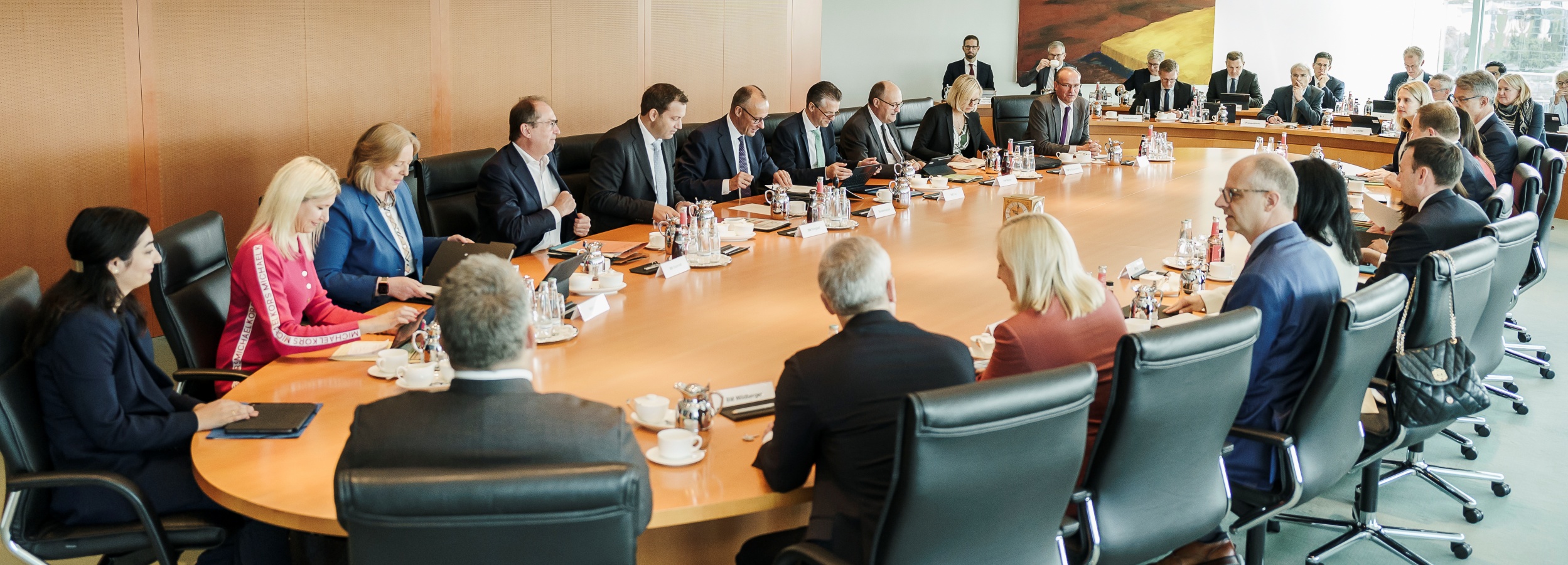 Das Bild zeigt die Gruppe von etwa 25 bis 30 Personen des Bundeskabinetts, die bei einer Kabinettssitzung um einen großen, ovalen Konferenztisch sitzen. Die Minister sind größtenteils in Businesskleidung gekleidet – Anzüge, Blazer und Blusen. Die Atmosphäre ist formell und konzentriert. An der Tischmitte befinden sich Getränke, Wasserkaraffen und kleine Teller mit Gebäck oder Süßigkeiten. Jeder Platz hat ein Namensschild. Einige der Teilnehmer scheinen aktiv an Diskussionen oder der Arbeit an Tablets und Dokumenten beteiligt zu sein. Hinter dem Haupt-Konferenztisch befindet sich ein weiteres kleines Gruppierung von Personen, die ebenfalls formell gekleidet sind und dem Geschehen am Haupttisch zu folgen scheinen. An der Wand hängt ein abstraktes Gemälde. Der Raum selbst ist modern und hell, mit hochwertigen Holzmöbeln und einem neutralen Farbschema. Die allgemeine Stimmung ist eine von konzentrierter Arbeit und formeller Interaktion. Es scheint sich um eine offizielle oder geschäftliche Sitzung zu handeln. Eine Handtasche ist auf einem Stuhl zu sehen. Die Beleuchtung ist hell und gleichmäßig, was auf einen gut ausgestatteten Konferenzraum hinweist.
