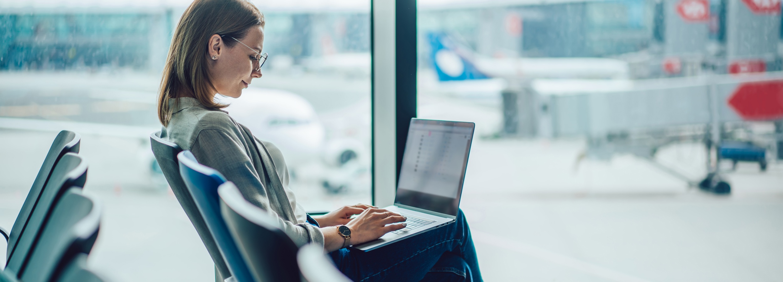 Das Bild zeigt eine Frau, die konzentriert an ihrem Laptop in einem Flughafen-Wartezimmer arbeitet.

Im Bild ist eine junge Frau mit braunen, schulterlangen Haaren zu sehen, die eine graue Bluse und Jeans trägt. Sie trägt eine Brille und eine Uhr am Handgelenk. Sie sitzt auf einem bequemen Stuhl in einem Flughafen-Wartezimmer und arbeitet an ihrem Laptop, der auf ihrem Schoß liegt. Ihre Hände tippen konzentriert auf die Tastatur. Ihr Blick ist auf den Bildschirm gerichtet und ihr Gesichtsausdruck wirkt fokussiert und konzentriert. Im Hintergrund ist durch ein großes Fenster die Rollbahn eines Flughafens mit Flugzeugen und Gepäckwagen unscharf zu sehen. Die Atmosphäre ist ruhig und konzentriert, obwohl der Ort ein geschäftiger Flughafen ist. Die Farben sind gedämpft, die Lichtverhältnisse hell und natürlich. Der gesamte Eindruck ist einer von konzentrierter Arbeit in einer öffentlichen, aber ruhigen Umgebung.