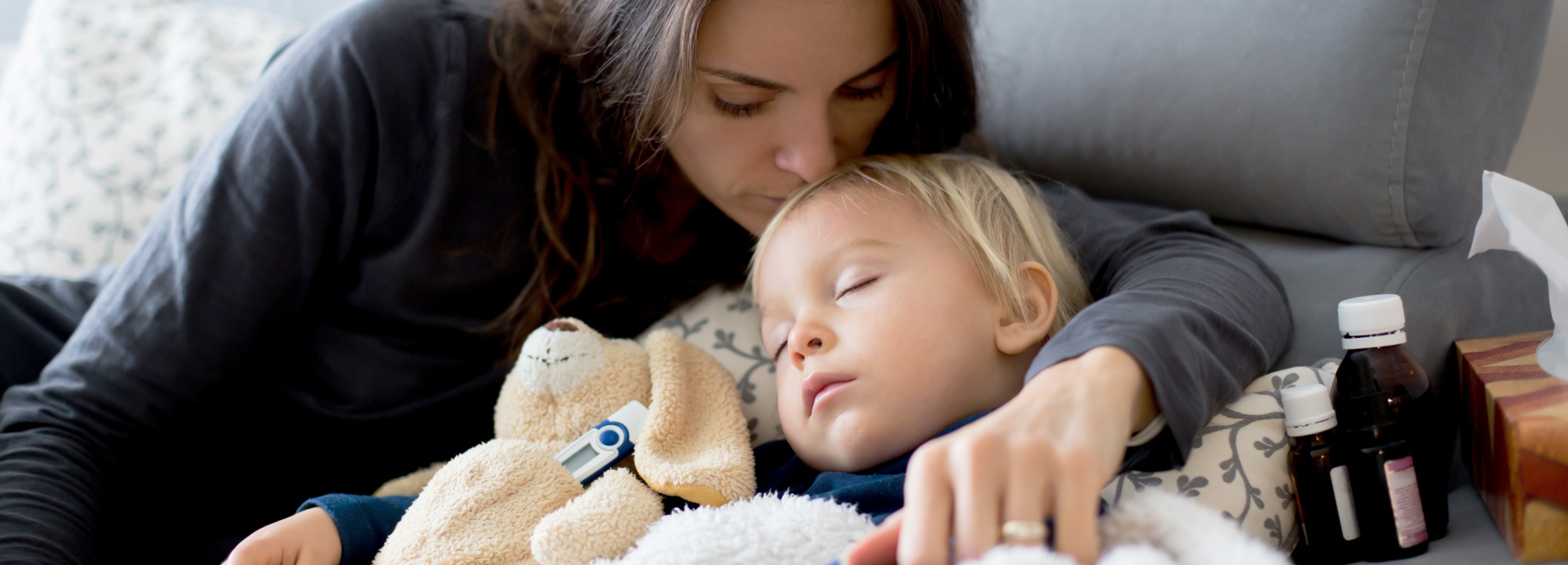Das Bild zeigt eine Mutter, die ihr krankes Kleinkind liebevoll auf der Couch tröstet.

Das Bild zeigt eine junge Frau mit langen, braunen Haaren, die ein kleines Kind mit blonden Haaren auf der Couch im Arm hält. Das Kind schläft und liegt eingekuschelt in eine Decke. Neben dem Kind liegt ein beigefarbenes Stofftier in Form eines Hundes, an dem ein digitales Fieberthermometer befestigt ist. Die Mutter beugt sich liebevoll über das Kind und legt ihr einen Kuss auf die Stirn. Ihr Gesichtsausdruck ist sanft und besorgt. Ihre Hand ruht tröstend auf dem Kind. Im Hintergrund sieht man einen Teil eines grauen Sofas und neben dem Kind stehen mehrere kleine Flaschen, die vermutlich Medikamente enthalten, sowie ein Paket Taschentücher. Die Atmosphäre des Bildes ist warm, ruhig und emotional, geprägt von Fürsorge und Zärtlichkeit. Die Farben sind gedämpft und natürlich. Das Gesamtbild vermittelt eine Stimmung von Geborgenheit und Fürsorge in einer Situation von Krankheit und Sorge um das Kind.