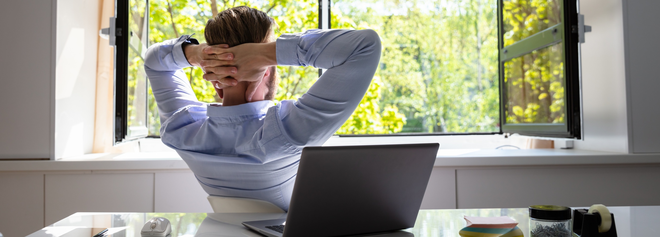 Ein Mann sitzt mit geschlossenen Augen und verschränkten Händen hinter dem Kopf entspannt vor einem geöffneten Fenster mit Blick auf grüne Bäume. Ein Laptop, eine Maus und Büromaterial liegen auf seinem Schreibtisch.